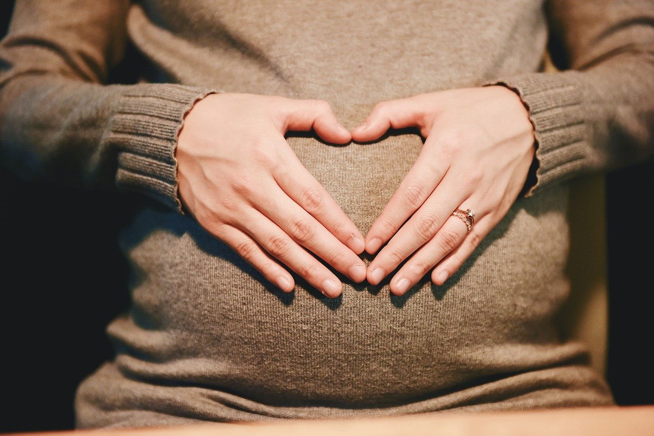 Gros plan d'une femme enceinte, mains formant un coeur sur le ventre, illustrant la douceur et la connexion pendant les cours de yoga prénatal et postnatal.